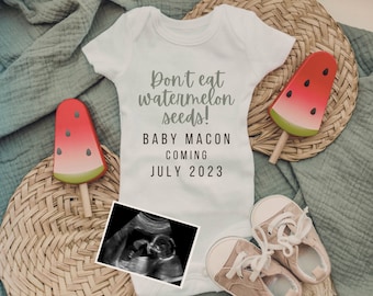 May include: A white baby onesie with the text "Don't eat watermelon seeds!" and "BABY MACON COMING JULY 2023". The image includes a sonogram, two watermelon-shaped wooden toys, and a pair of baby shoes.
