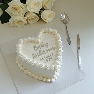 May include: A white heart-shaped cake with white frosting and the text "Baby Anderson October 2024" written in gray frosting. A bouquet of white roses is in the background.