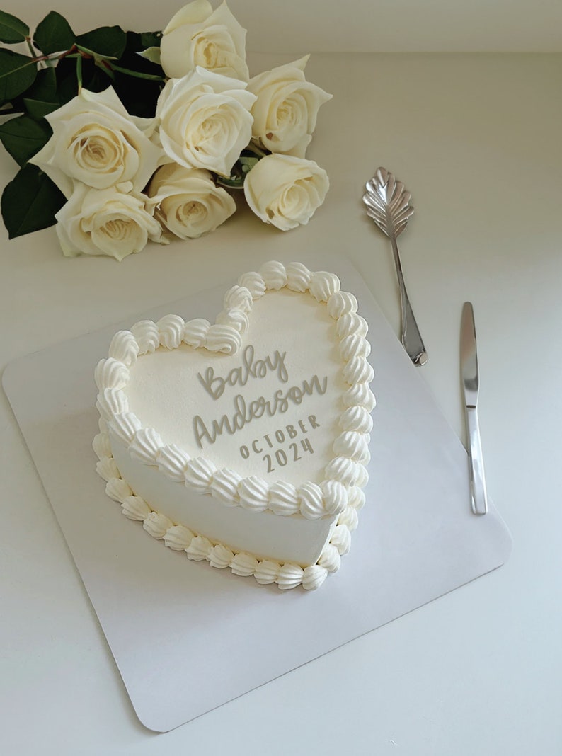 May include: A white heart-shaped cake with white frosting and the text "Baby Anderson October 2024" written in gray frosting. A bouquet of white roses is in the background.