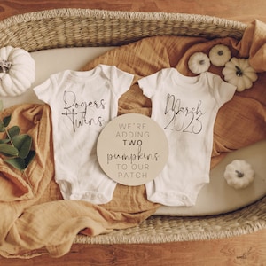 Puede incluir: Dos bodies blancos con el texto "Rogers Twins" y "Marzo 2023" en ellos. Un letrero de madera con el texto "We're adding two pumpkins to our patch" está en el centro de la imagen. Los bodies y el letrero están en una cesta de mimbre con calabazas blancas y una planta verde.