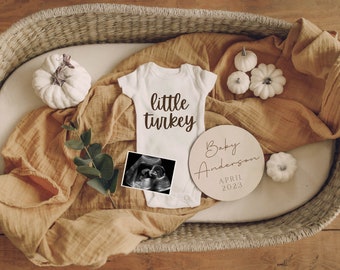 May include: A white onesie with the text "little turkey" on it, a wooden sign that says "Baby Anderson April 2023", and a small ultrasound image in a wicker basket with a tan blanket and white pumpkins.