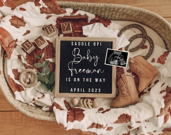 May include: A black letter board with the text "Saddle Up! Baby Freeman is on the way April 2023".  The letter board is surrounded by a brown and white cow print blanket, wooden blocks spelling "BABY", a small white onesie, two brown cowboy boots, and a brown horseshoe.