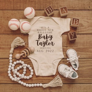 Könnte beinhalten: Ein weißer Strampler mit dem Text "It's a whole new ballgame Baby Taylor arriving Aug 2022" darauf. Der Strampler ist umgeben von Baseballs, Holzklötzen, die "BABY" buchstabieren, einer Holzrassel und einer weißen Perlenkette.