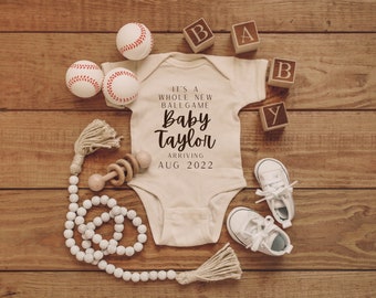 Puede incluir: Un body blanco con el texto "It's a whole new ballgame Baby Taylor arriving Aug 2022" en él. El body está rodeado de pelotas de béisbol, bloques de madera que deletrean "BABY", un sonajero de madera y una guirnalda de cuentas blancas.