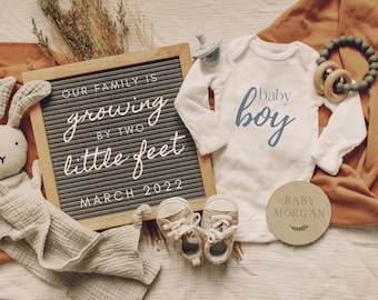May include: A baby announcement with a letter board that reads "Our family is growing by two little feet March 2022". There is a white onesie with the text "baby boy" in blue script, a pair of baby shoes, a wooden circle with the text "Baby Morgan", and a stuffed bunny.