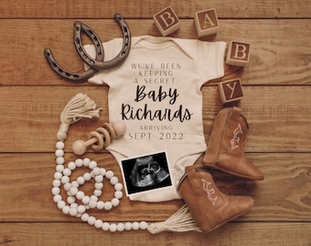 May include: A white onesie with the text "We've been keeping a secret... Baby Richards Arriving Sept 2022" and a black and white ultrasound image. The onesie is surrounded by wooden blocks that spell "BABY", two brown cowboy boots, a wooden rattle, a string of white beads, and two brown horseshoes.