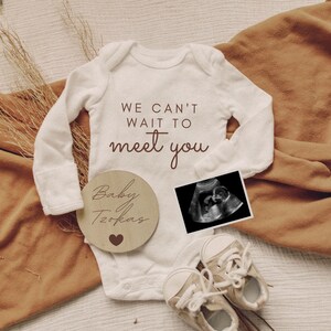 K&ouml;nnte beinhalten: Ein wei&szlig;er Strampler mit dem Text "We can't wait to meet you". Ein Holz-Kreis mit dem Text "Baby [name]" und einem Herz. Ein Schwarzwei&szlig;-Ultraschallbild. Ein Paar wei&szlig;e Babyschuhe.