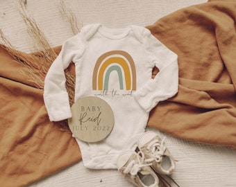May include: A white baby onesie with a rainbow graphic and the text "worth the wait". A wooden circle with the text "Baby Reid July 2022" is next to the onesie.
