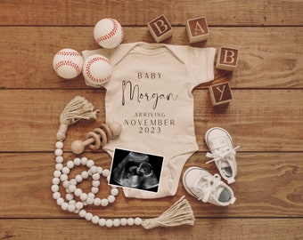 Puede incluir: Un body blanco con el texto "Baby Morgan Arriving November 2023". El body está rodeado de pelotas de béisbol, bloques de madera que deletrean "BABY", un sonajero de madera, una cadena de cuentas blancas y una imagen de sonograma.