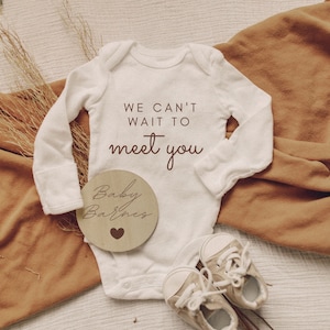 K&ouml;nnte beinhalten: Wei&szlig;er Baby-Strampler mit dem Text "We can't wait to meet you". Ein Holz-Kreis mit dem Text "Baby Barnes" und einem kleinen Herz liegt neben dem Strampler.