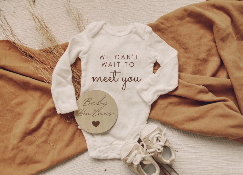 K&ouml;nnte beinhalten: Wei&szlig;er Baby-Strampler mit dem Text "We can't wait to meet you". Ein Holz-Kreis mit dem Text "Baby Barnes" und einem kleinen Herz liegt neben dem Strampler.
