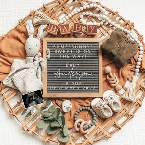 May include: A letter board with the text "Some bunny sweet is on the way! Baby Anderson is due December 2023". The board is surrounded by baby items, including a stuffed bunny, a rattle, a pair of baby shoes, and a wooden bead garland.