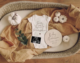 Puede incluir: Una escena de anuncio de bebé con un body blanco con la frase "Our Little Pumpkin", una ecografía y un disco de madera con "Baby Anderson January 2023". La escena incluye pequeñas calabazas blancas y una manta color mostaza.
