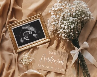 May include: A tan fabric background with a gold picture frame containing a black and white ultrasound image. A bouquet of white baby's breath flowers with a tan ribbon is on the right side of the image. A tan card with the text "BABY Anderson JANUARY 2026" is in the center of the image.