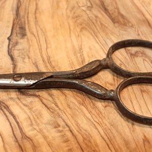 May include: Antique metal scissors with oval finger holes and a pointed blade. The scissors have a rusty, aged appearance, suggesting they are vintage or well-used. The scissors are lying on a wooden surface.