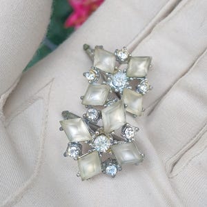 May include: A pair of vintage silver-toned earrings. Each earring features a central cluster of clear, round rhinestones surrounded by pale, square-cut stones. Additional smaller rhinestones are interspersed throughout the design.