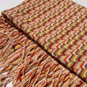 May include: A folded, fringed scarf with a chevron pattern. The scarf features a mix of colors including beige, orange, yellow, and brown. The fringe is a blend of orange, beige, and yellow threads.