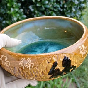 May include: A handcrafted ceramic bowl with a textured, carved exterior and a glossy, teal interior. The rim features embossed Chinese characters and a dark, abstract design. The bowl is approximately 10 inches in diameter.