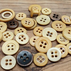 May include: A collection of various wooden buttons in shades of beige, brown, and black. The buttons vary in size and shape, including round and ring-shaped designs, with four-hole and two-hole styles. The buttons are arranged on a wooden surface.