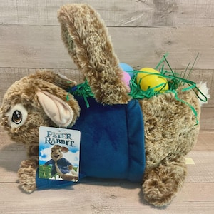 May include: A plush Easter bunny basket with a blue body and brown fur. The basket is filled with green faux grass and colorful Easter eggs. A tag featuring Peter Rabbit is attached.