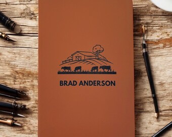 May include: A brown notebook with a farm scene illustration and the name "BRAD ANDERSON" printed in black. The illustration features a barn, tree, and cows. The notebook is on a wooden surface.