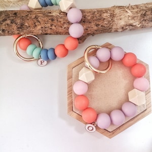 Könnte beinhalten: Zwei bunte Schlüsselanhänger aus Silikonperlen mit Holzperlen und goldenen Schlüsselringen. Der Schlüsselanhänger links hat blaue, grüne und orange Perlen mit einer Holzperle in Form eines Quadrats. Der Schlüsselanhänger rechts hat lila, rosa und orange Perlen mit einer Holzperle in Form eines Quadrats. Beide Schlüsselanhänger haben einen goldenen Anhänger mit einem Buchstaben darauf.