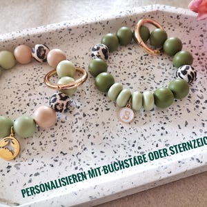 Könnte beinhalten: Zwei Armbänder mit grünen, beigen und braunen Perlen. Ein Armband hat einen goldenen Anhänger mit dem Buchstaben S. Das andere Armband hat einen goldenen Anhänger mit einem Blattdesign. Die Armbänder befinden sich auf einem weißen Tablett mit dem Text "PERSONALISIEREN MIT BUCHSTABE ODER STERNZEICHEN".