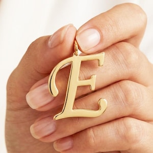 Può includere: Un ciondolo in metallo color oro lucido a forma di lettera maiuscola E. La lettera presenta un carattere stilizzato ispirato al corsivo con superfici lisce e riflettenti e un piccolo anello circolare sulla parte superiore per appenderlo.