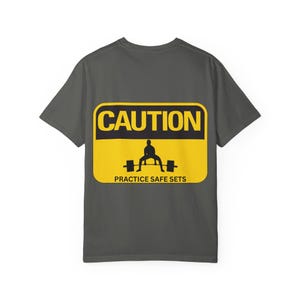 Könnte beinhalten: Dunkelgraues T-Shirt mit gelb-schwarzer Grafik. Die Grafik zeigt ein Warnschild mit dem Wort "CAUTION" über einer Silhouette einer Person beim Gewichtheben und dem Text "PRACTICE SAFE SETS" darunter.