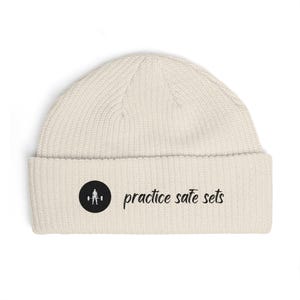 Könnte beinhalten: Cremefarbene Beanie-Mütze mit Umschlag. Auf der Mütze befindet sich ein schwarzes rundes Emblem mit einer Gewichtheber-Silhouette und dem Schriftzug "practice safe sets" in Schwarz.