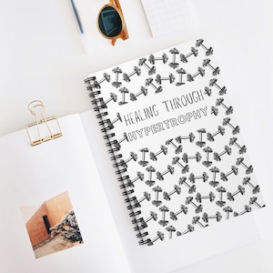 Könnte beinhalten: Ein weißes Spiralnotizbuch mit einem Muster aus schwarzen Hanteln und dem Text "HEALING THROUGH HYPERTROPHY". Das Notizbuch liegt offen auf einer weißen Oberfläche. Eine goldene Büroklammer und eine Sonnenbrille sind ebenfalls zu sehen.