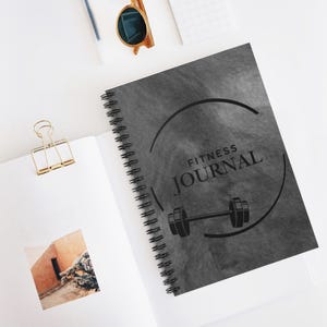 May include: A spiral-bound fitness journal with a gray textured cover. The cover features the words "FITNESS JOURNAL" in a circular design, along with a barbell graphic. The journal is open, revealing blank pages and a photo.