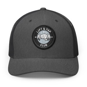 May include: A charcoal gray trucker hat with a black mesh back. The front of the hat features a circular patch with the words "LIFT & YAP CLUB" surrounding a weightlifting design with a fist. The hat has a curved brim.
