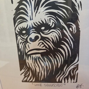 Può includere: Stampa in bianco e nero del volto di una creatura simile a Bigfoot. L'opera presenta linee audaci e un aspetto strutturato, con la frase "GONE SQUATCHIN'" in basso. L'immagine è incorniciata in un bordo bianco.