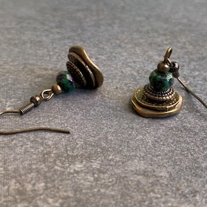 Könnte beinhalten: Ein Paar grüne und bronzene Ohrringe mit einem geschichteten Design. Die Ohrringe haben eine grüne Steinperle und ein geschichtetes Bronze-Design.