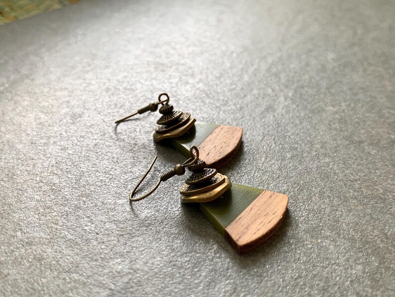 May include: A pair of earrings with a green and brown wood design. The earrings feature a gold-toned metal charm with a layered design.