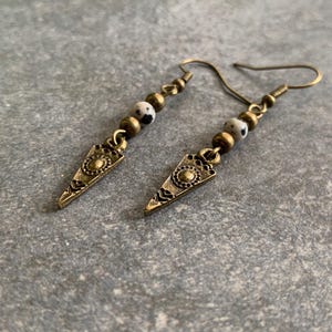 Könnte beinhalten: Ein Paar Ohrringe mit goldfarbenem Finish. Jeder Ohrring hat eine kleine, weiß-schwarz gesprenkelte Perle und einen dreieckigen Anhänger mit filigranen Details.