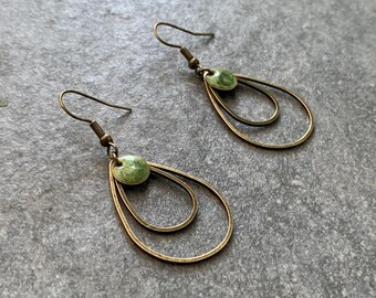 May include: A pair of brass-toned earrings with a double teardrop design. Each earring features a small, round, green bead at the top. The earrings have a simple hook closure and are displayed on a textured gray surface.