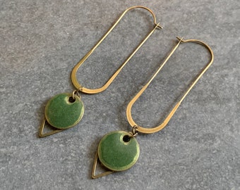May include: A pair of gold-toned earrings with a modern design. Each earring features a long, curved bar and a green circular pendant with a triangular accent. The earrings are displayed on a gray surface.