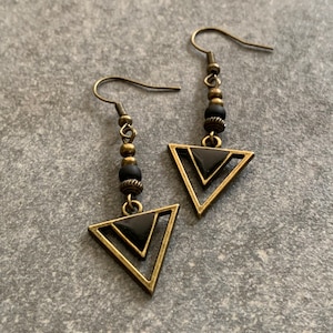 Könnte beinhalten: Ein Paar goldene Dreiecksohrringe mit schwarzen Akzenten. Die Ohrringe haben ein geometrisches Design und hängen an einem Haken.