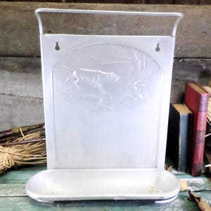 May include: A vintage, silver-coloured metal wall-mounted organiser with a hunting scene embossed on the front. It features a small shelf at the bottom and a handle at the top. The organiser is likely used for storing small items.