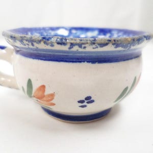Puede incluir: Una taza de té de cerámica blanca con un borde azul y un patrón floral. La taza tiene un asa y está decorada con flores naranjas, verdes y azules.