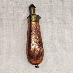 May include: An antique copper powder flask with brass detailing. The flask has a teardrop shape and a textured surface. The top and spout are brass, and there is a small ring at the base. Likely from the 1800s.