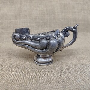 Peut inclure: Une lampe à huile en métal argenté présentant un corps incurvé et allongé avec des volutes en relief, une série de petits points sur le côté, une base circulaire sur pied, ainsi qu'une anse avec un détail enroulé et une large ouverture.