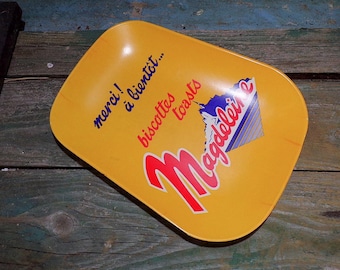 May include: Yellow serving tray with the text "merci! à bientôt... biscottes toasts Madeleine" in red and blue. The tray has a rounded rectangular shape and a graphic of a mountain and stripes.