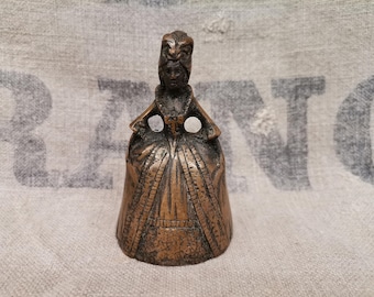May include: A bronze-colored bell shaped like a woman in a long dress. The bell has a detailed design, including the woman's face, hair, and clothing. Two holes are visible near the top. The bell is likely a decorative item.
