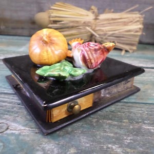 Puede incluir: Una caja de baratijas negra y rectangular con un pequeño cajón y un pomo de latón. La parte superior de la caja presenta verduras de cerámica, incluyendo una cebolla amarilla, una cebolla roja y blanca y hojas verdes. La caja está sobre una superficie de madera envejecida.