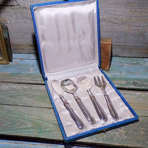 Może przedstawiać: Zestaw czterech srebrnych sztućców do serwowania w niebieskim pudełku wyłożonym aksamitem. Sztućce obejmują nóż do masła, łyżkę, widelec i małą łyżkę do serwowania.