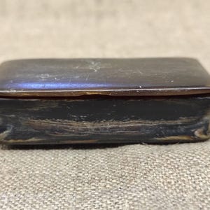 Puede incluir: Una pequeña caja rectangular de color marrón oscuro con una tapa lisa y pulida y una base ligeramente cóncava. La superficie presenta patrones de vetas naturales y arremolinados en tonos marrón oscuro y negro, con un diseño grabado.