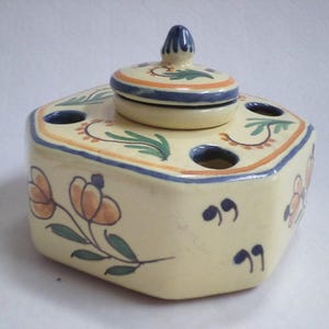 Pode incluir: Um tinteiro hexagonal de cerâmica com tampa, apresentando um design floral em tons de azul, verde, laranja e amarelo. O tinteiro tem seis orifícios ao redor da parte superior para canetas ou lápis.